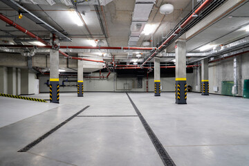 Fototapeta premium Modern underground parking garage with concrete columns, red fire sprinkler pipes, ventilation ducts and drainage channels in a newly constructed residential building