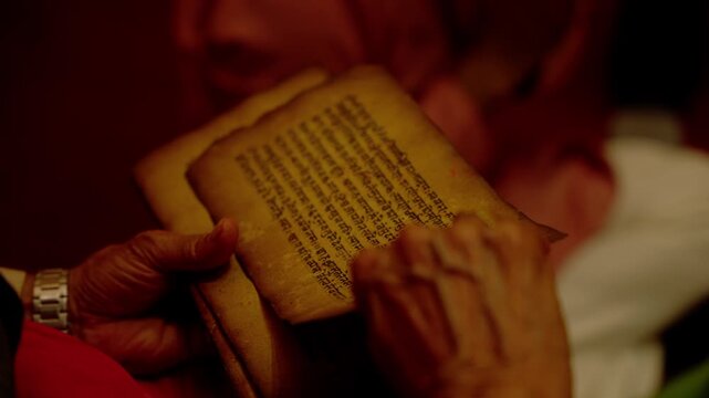 Intimate close up of two aged hands holding open pages of ancient handwritten religious text illuminated by warm indoor lighting