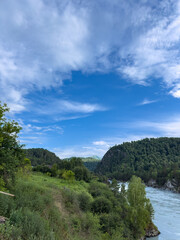 Fototapeta premium A Serene Waterway Surrounded by Lush Greenery and Majestic Hills Under a Blue Sky with Fluffy Clouds