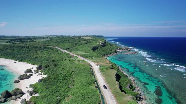 断崖沿いの海岸道路と白い砂浜｜宮古島・沖縄のエメラルドグリーンの海を望む絶景ドライブ空撮/Aerial view of scenic coastal road along limestone cliffs with turquoise ocean and white sand beach, Miyako Island, Okinawa, Japan