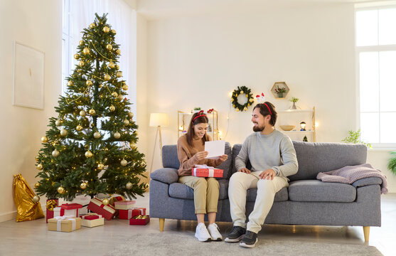 Christmas couple exchanging gift at cozy home. They sit by festive tree, sharing present and reading greeting card, smiling with love and surprise in decorated room. Celebration and giving.