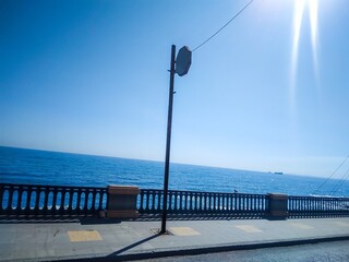 Fototapeta premium Tall metal utility pole stands against clear blue sky on seaside promenade featuring protective stone railing overlooking deep ocean waters under bright summer sun rays