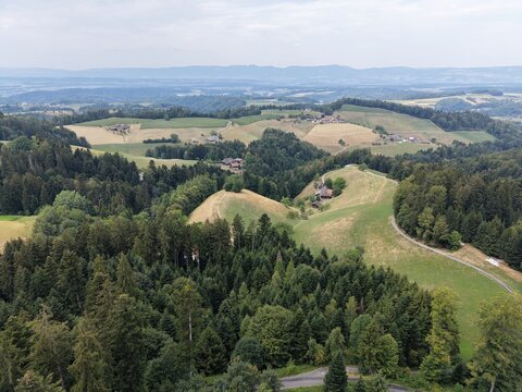 Aerial view of the Emmental region featuring rolling green hills, dense forests, scattered traditional farmhouses, and winding country roads in Ruderswil, Canton of Bern, Switzerland.