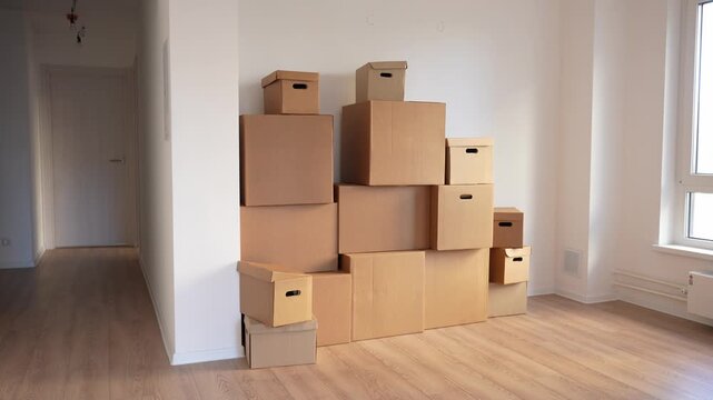 Stacked cardboard boxes fill a corner. Boxes form a tall, unstable pyramid. White walls and wooden floor frame the scene. Hallway leads to a distant door. Sunlight streams through a window