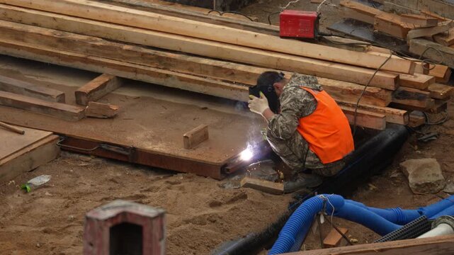 Worker in orange vest welds metal beams. Bright sparks fly from his welding torch. Wooden planks and construction materials surround him. Dirt ground holds tools and bricks