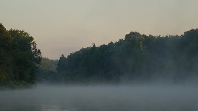Misty lake surface shimmers with morning light. Dense forest looms behind the water. Soft golden hues glow in the sky. Perfect for tranquil nature shots. Early dawn hours capture this peaceful moment