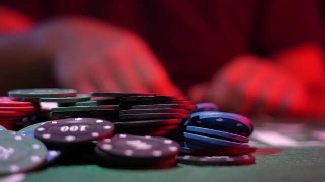 Colorful poker chips stack on green felt. Red background hints at casino atmosphere. Shallow focus highlights chip details. Blurred hands suggest active game