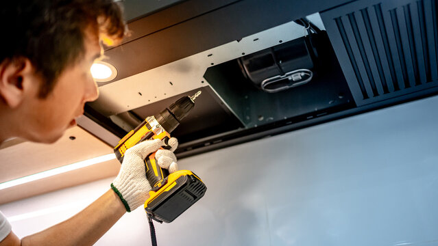 Asian male technician using electric screwdriver fixing extractor motor in cooker hood in modern kitchen. Maintenance worker man repairing exhaust fan in kitchen hood. Home appliance service