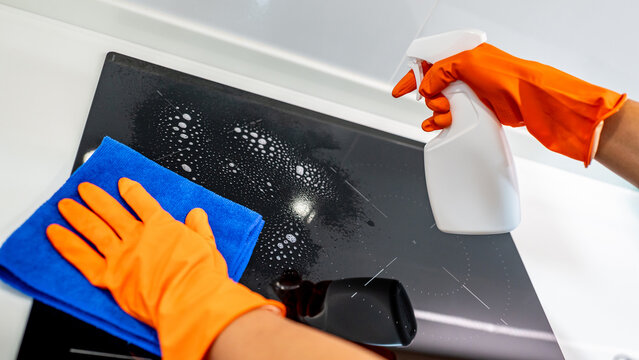 Housekeeper with orange rubber gloves spraying soapy detergent on induction cooktop wiping blue microfiber cloth on kitchen counter. Home hygiene maintenance. Cleaning tools for housework chores