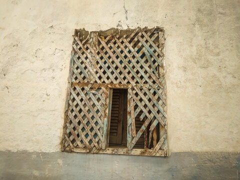 Weathered wooden lattice window screen with peeling blue paint covers a rustic aged stucco wall of an old historic Mediterranean house during a sunny day