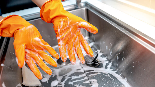 Housekeeper showing hands with orange rubber glove full with soap suds while scrubbing on kitchen sink surface. Cleaning stainless steel sink for hygiene. Housework chores in the kitchen