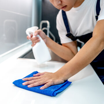 Asian male housekeeper holding detergent spray bottle while wiping blue microfiber cloth on modern white countertop in domestic kitchen. Home hygiene maintenance. Cleaning house service