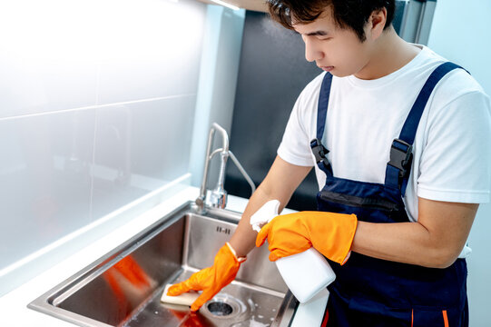 Asian male housekeeper wearing orange rubber gloves holding detergent spray bottle while scrubbing stainless steel kitchen sink with sponge. Home hygiene maintenance service