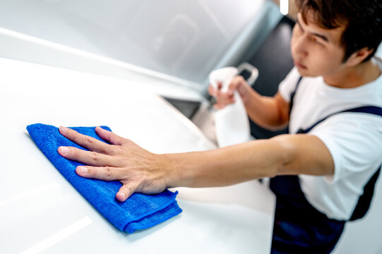 Asian male housekeeper holding detergent spray bottle while wiping blue microfiber cloth on modern white countertop in domestic kitchen. Home hygiene maintenance. Cleaning house service