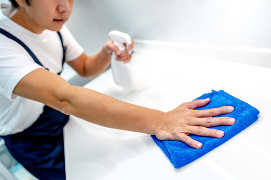 Asian male housekeeper holding detergent spray bottle while wiping blue microfiber cloth on modern white countertop in domestic kitchen. Home hygiene maintenance. Cleaning house service