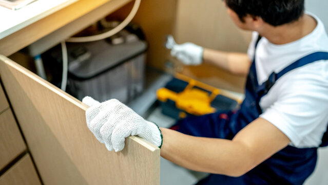 Male plumber with work tools opening wooden cabinet checking grease trap box and pipe connection under the kitchen sink. Handyman prepares to fix the plumbing system.