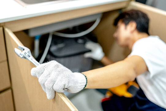 Asian male plumber with glove and adjustable wrench in his hand opening wooden cabinet checking grease trap box and pipe system under the kitchen sink. Handyman fixing plumbing system in the kitchen.
