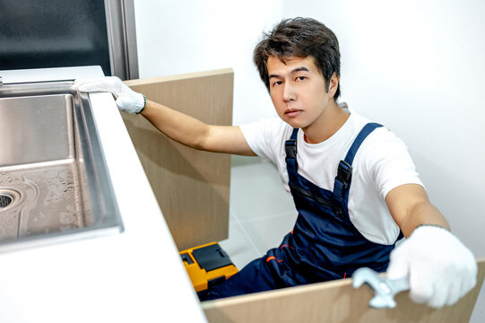 Confident asian male plumber with glove and adjustable wrench in his hand opening wooden cabinet checking pipe system under the stainless steel sink. Handyman looking at camera in the kitchen.