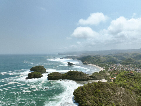 Aerial view of Pantai Kasap featuring rocky islets, crashing waves, lush green coastal cliffs, and a small village nestled along the shoreline in Kecamatan Pringkuku, Jawa Timur, Indonesia.