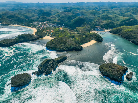 Aerial view of Pantai Kasap featuring rugged rocky islets, turquoise waves crashing against the shore, lush green hills, and sandy beaches in Kecamatan Pringkuku, Jawa Timur, Indonesia.