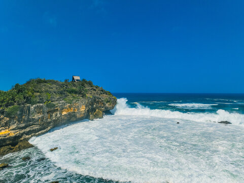 Aerial view of Pantai Kasap featuring a rocky cliff with a small wooden structure, surrounded by crashing white waves and deep blue ocean in Kecamatan Pringkuku, Jawa Timur, Indonesia.