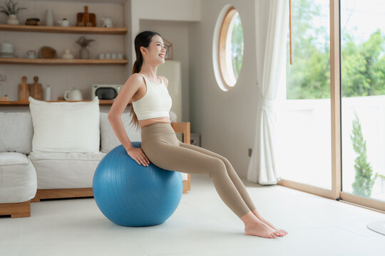 Asian woman in beige activewear sits on a blue stability ball with hands on the ball and smiles while looking toward the window.
