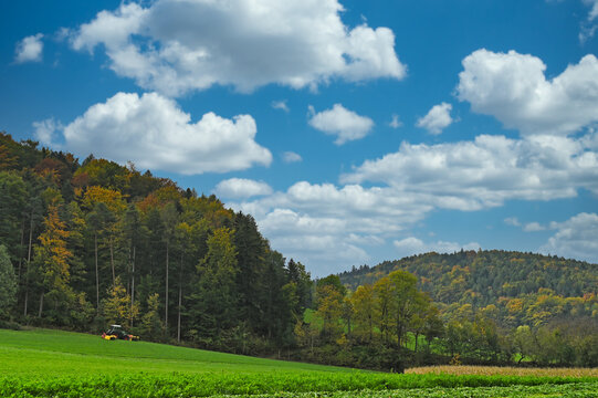 Modern tractor mowing green grass for hay next to a colorful autumn forest and rolling hills in Lower Austria