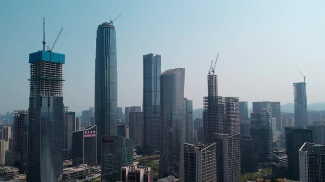 A modern urban skyline featuring multiple skyscrapers, several of which are under construction with cranes visible against a clear daytime sky.
