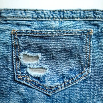 close-up of a blue denim back pocket with two frayed horizontal rips, contrasting orange stitching, visible white pocket lining and a casual worn rugged feel