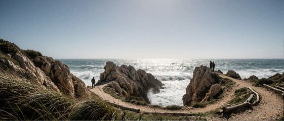 Fototapeta premium Panoramic view of rugged coastal cliffs with people walking on a sandy path overlooking the ocean