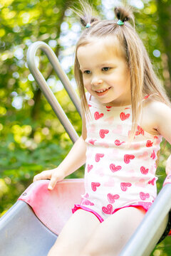 Young girl with long hair wearing a heart-patterned tank top slides down a playground slide surrounded by green trees and sunlight in a park setting