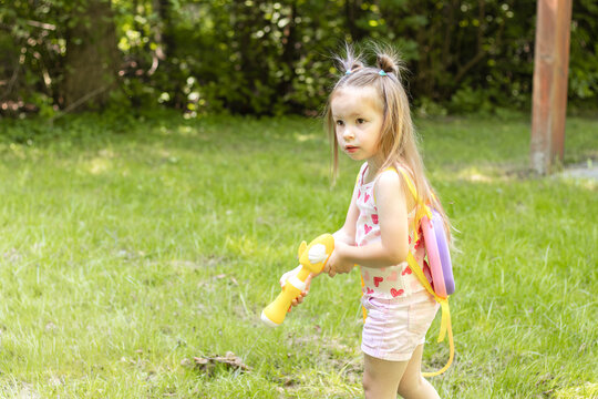 Young girl with long hair wearing a pink and white outfit holds a yellow toy water gun while playing in a grassy area surrounded by trees