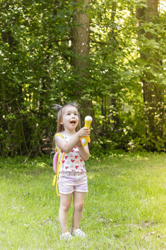 Young girl with brown hair wearing a heart-patterned shirt and shorts plays with a toy in a green forested area, sunlight filtering through the trees