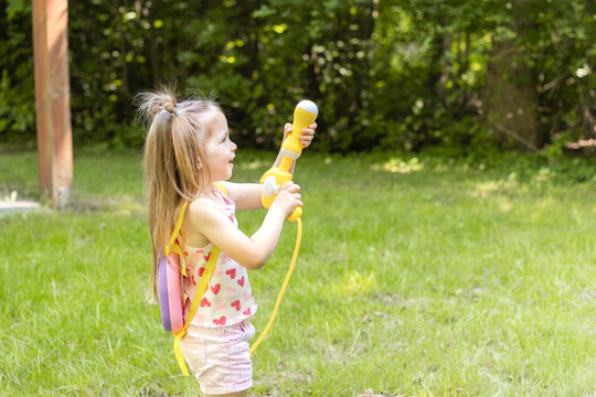 Young girl with long hair wearing a pink heart-patterned tank top and shorts plays with a colorful water toy in a grassy backyard under bright sunlight