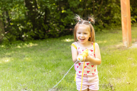 Young girl with pigtails wearing a pink tank top and shorts holds a water toy while playing in a green backyard with trees and sunlight filtering through