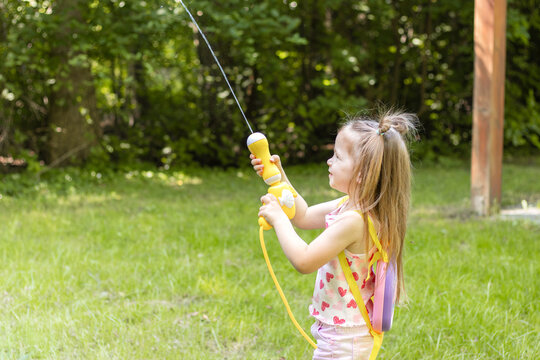 Young girl with long hair wearing pink outfit holds a toy fishing rod while standing on green grass in a sunny outdoor setting surrounded by trees