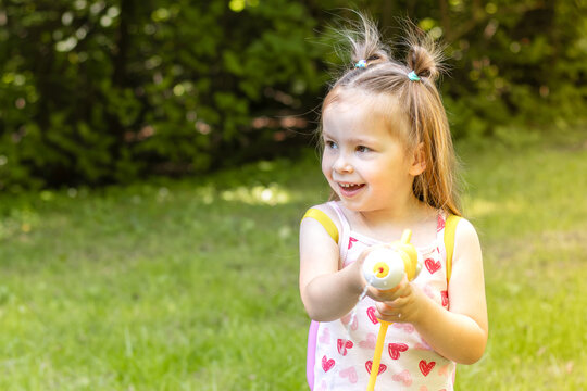 Young girl with playful hairstyle wearing a pink heart-patterned tank top holds a water gun while standing on green grass in a sunny outdoor setting