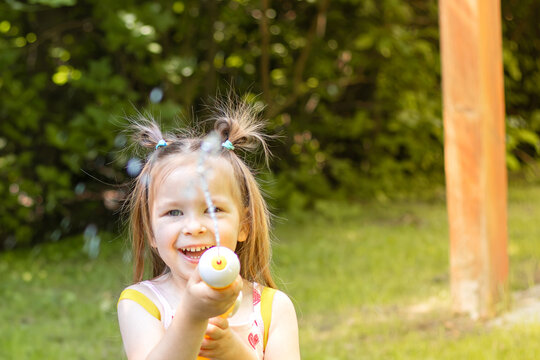 Young girl with pigtails playfully sprays water from a toy water gun in a sunny backyard, surrounded by greenery and wooden posts in the background