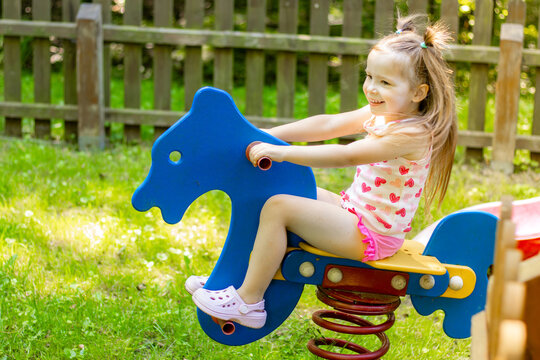 Young girl with light brown hair wearing a pink tank top and shorts enjoys riding a blue horse-shaped spring toy in a grassy playground area surrounded by wooden fencing