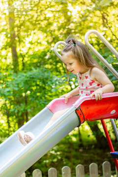 Young girl with brown hair wearing a pink dress slides down a playground slide surrounded by green trees and sunlight in a park setting
