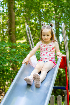Young girl with light brown hair wearing a pink heart-patterned tank top slides down a playground slide surrounded by green trees in a sunny park setting