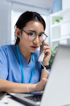 women in blue scrubs answering telephone while using laptop in medical office, representing patient support, telehealth service and healthcare communication.
