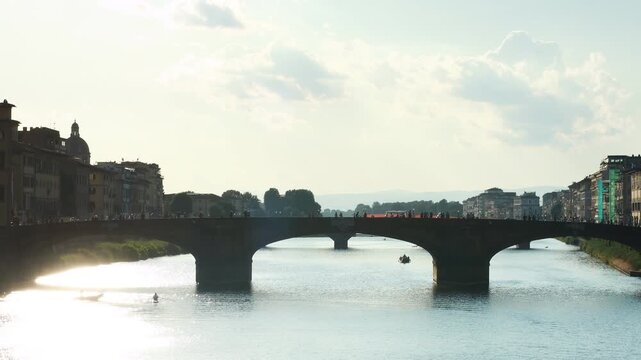 Ponte alla Carraia over the Arno river in Florence, Italy. Static shot of the bridge in backlight with the San Frediano dome, riverside palazzi and rowing boats on the water in summer
