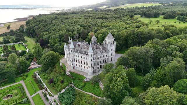 Wide angle panoramic aerial drone shot of Dunrobin Castle surrounded by green woodland trees, formal French landscape gardens, and coastal sea bay in Scottish Highlands