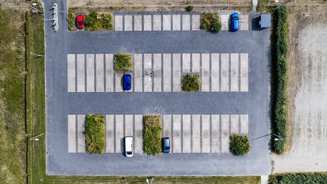 Aerial view of a paved parking lot with marked spaces, scattered cars, and small green patches of vegetation in Delft, South Holland, Netherlands.