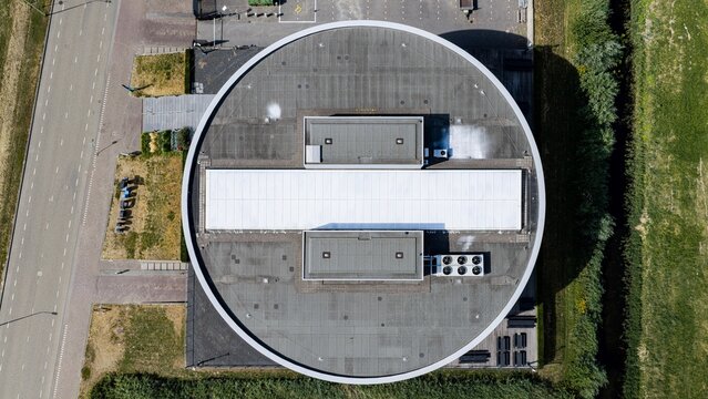 Aerial view of a modern circular building with a flat roof and HVAC units, situated next to a road and green fields in Delft, South Holland, Netherlands.
