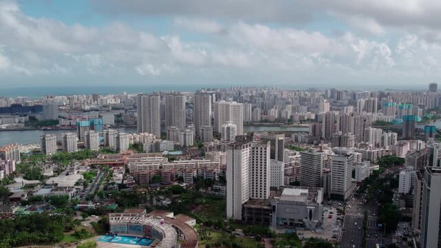 A daytime aerial panorama of a dense coastal urban area, featuring numerous high-rise buildings, water bodies, and green spaces under a partly cloudy sky.