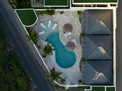 Aerial view of a modern villa with a kidney-shaped swimming pool, lounge chairs, straw umbrellas, and lush palm trees in Kecamatan Kuta Selatan, Bali, Indonesia.