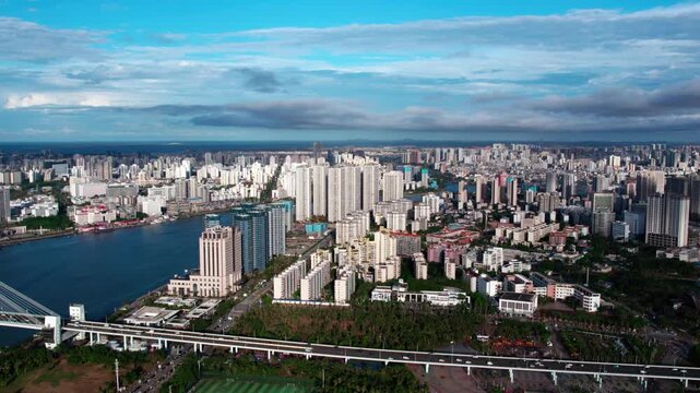 An aerial panorama of a dense urban area with a river, coastal bridge, and high-rise buildings under a blue sky.