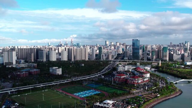This aerial shot shows a modern urban cityscape with high-rise buildings, an overpass, green spaces, and a river under a partly cloudy sky.